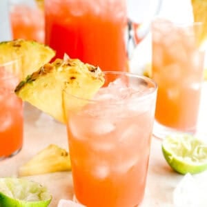 Tall glass pitcher of bright red fruit punch filled with ice, surrounded by glasses of punch topped with fresh pineapple wedges and scattered ice and lime on the table.