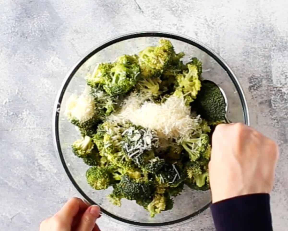 Hands mixing fresh broccoli florets with grated Parmesan cheese in a glass bowl before roasting.