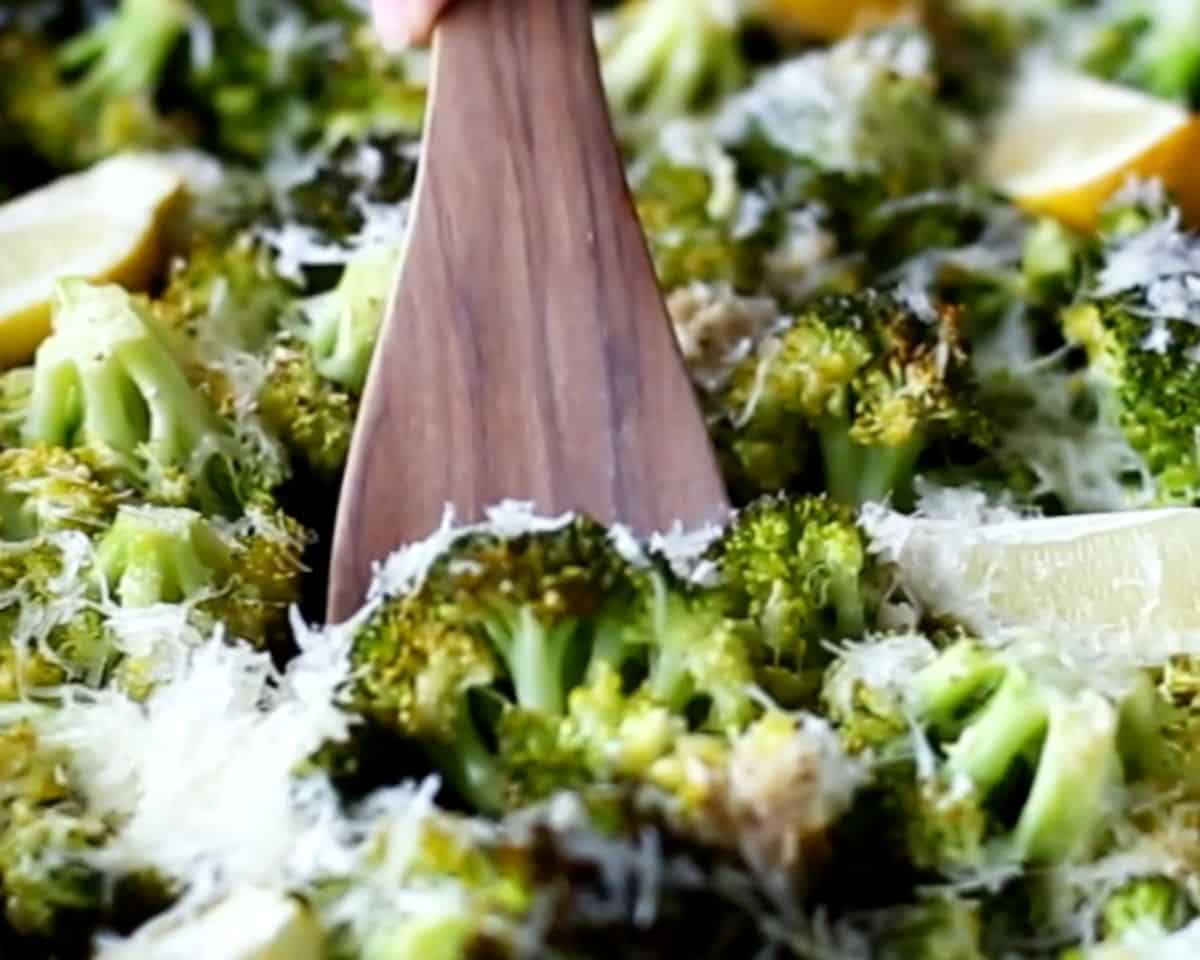 Wooden spoon stirring hot roasted broccoli with melted Parmesan and roasted lemon wedges on a sheet pan.