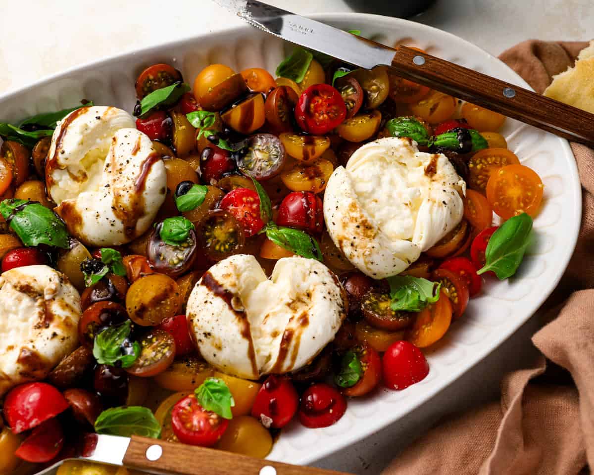 Burrata caprese with a knife cutting into the cheese, revealing the creamy center alongside tomatoes, basil, and balsamic drizzle.