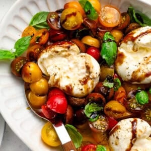 Close-up of burrata caprese with creamy burrata torn open, mixed cherry tomatoes, fresh basil, and balsamic reduction on a white serving platter.