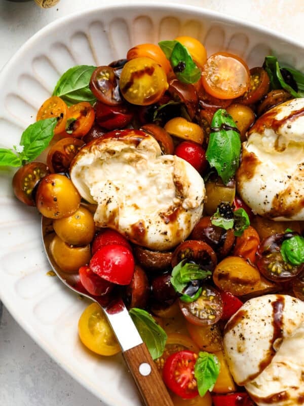 Close-up of burrata caprese with creamy burrata torn open, mixed cherry tomatoes, fresh basil, and balsamic reduction on a white serving platter.