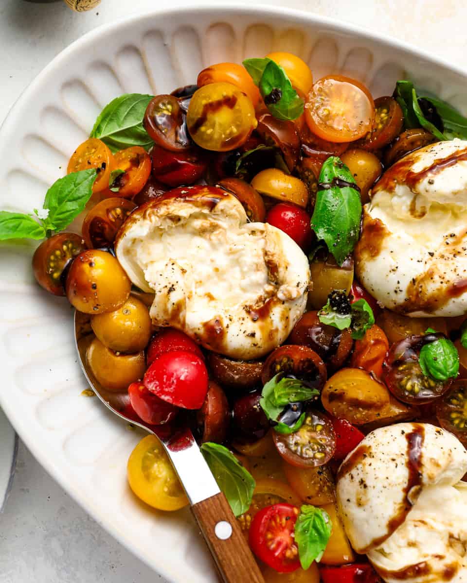 Close-up of burrata caprese with creamy burrata torn open, mixed cherry tomatoes, fresh basil, and balsamic reduction on a white serving platter.