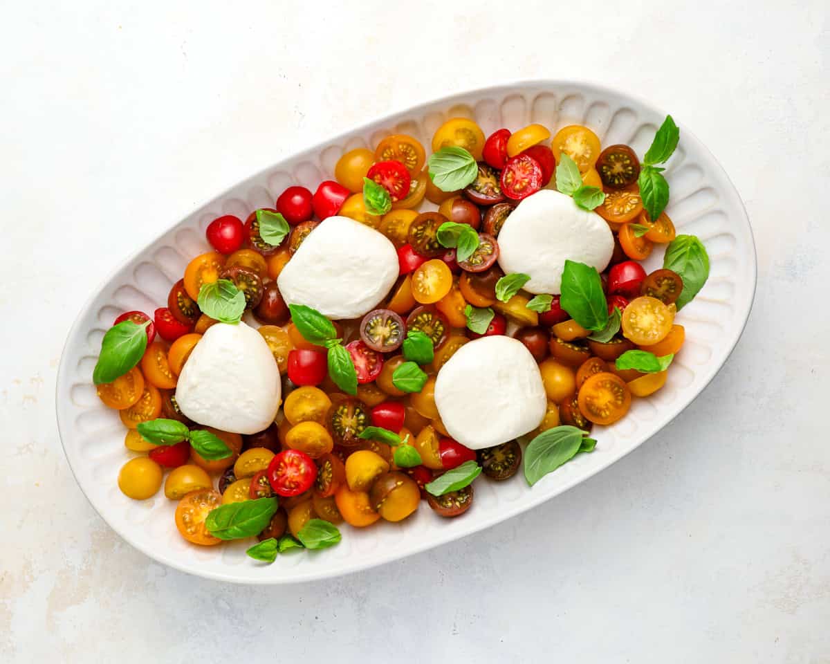 Burrata balls placed over tomatoes with fresh basil leaves scattered on top on a white oval platter.
