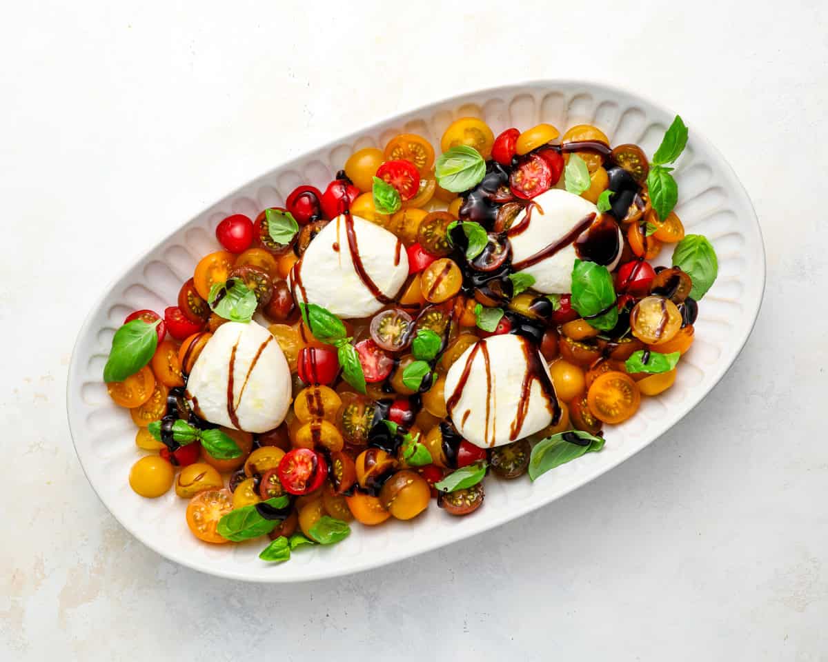 Burrata caprese salad drizzled with balsamic reduction, showing colorful tomatoes, basil, and whole burrata balls.