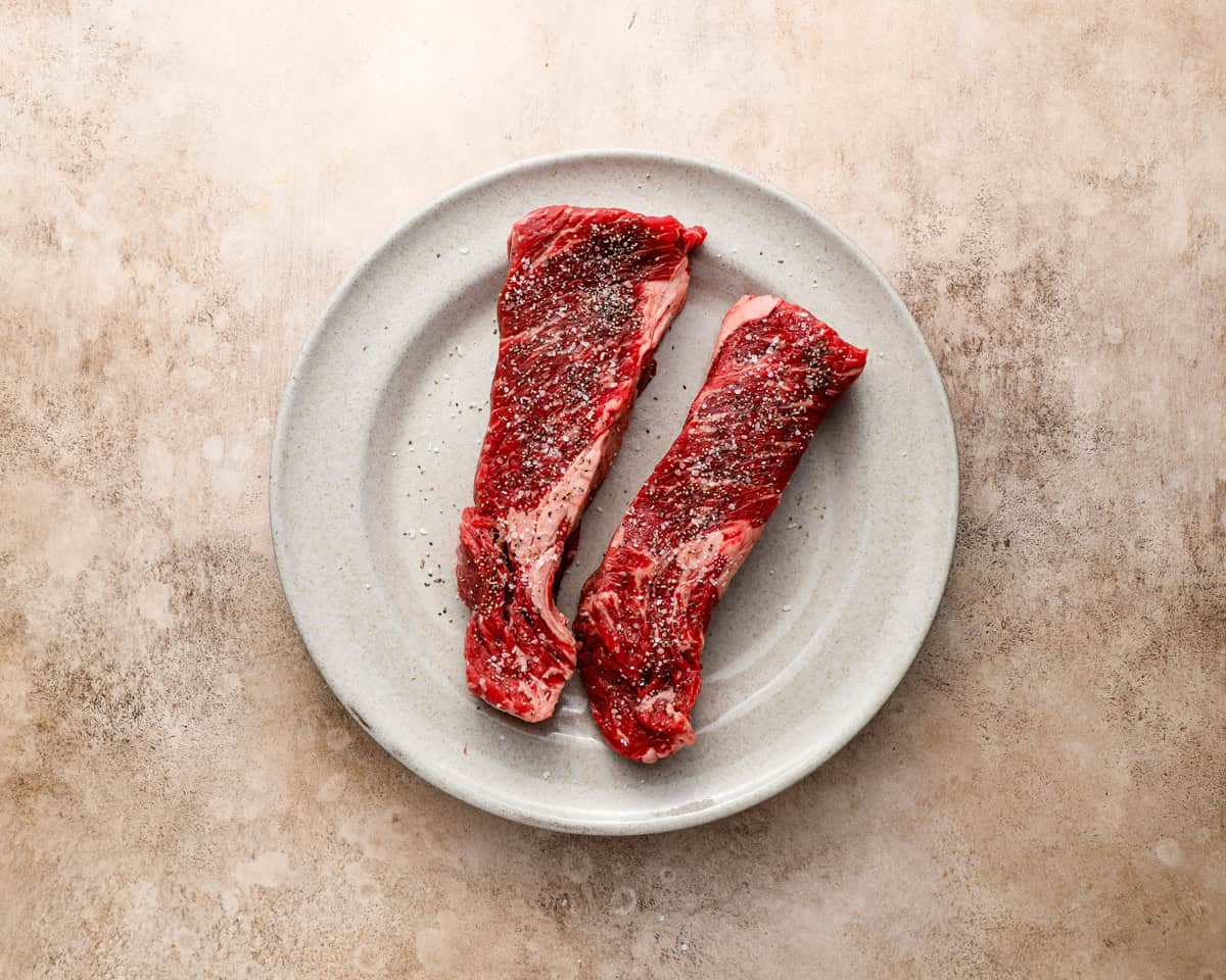 Raw Denver steaks seasoned with salt and black pepper on a white plate before cooking