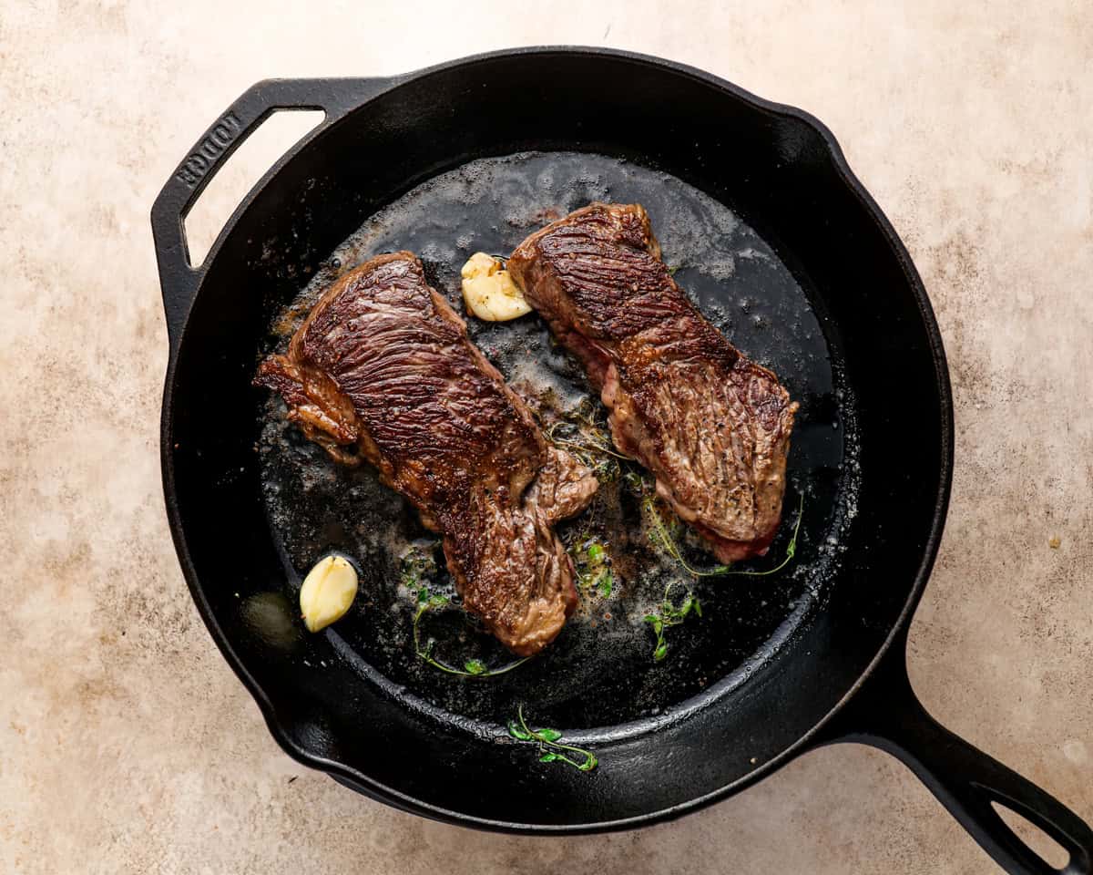 Denver steaks searing in a cast iron skillet with garlic cloves and thyme in melted butter