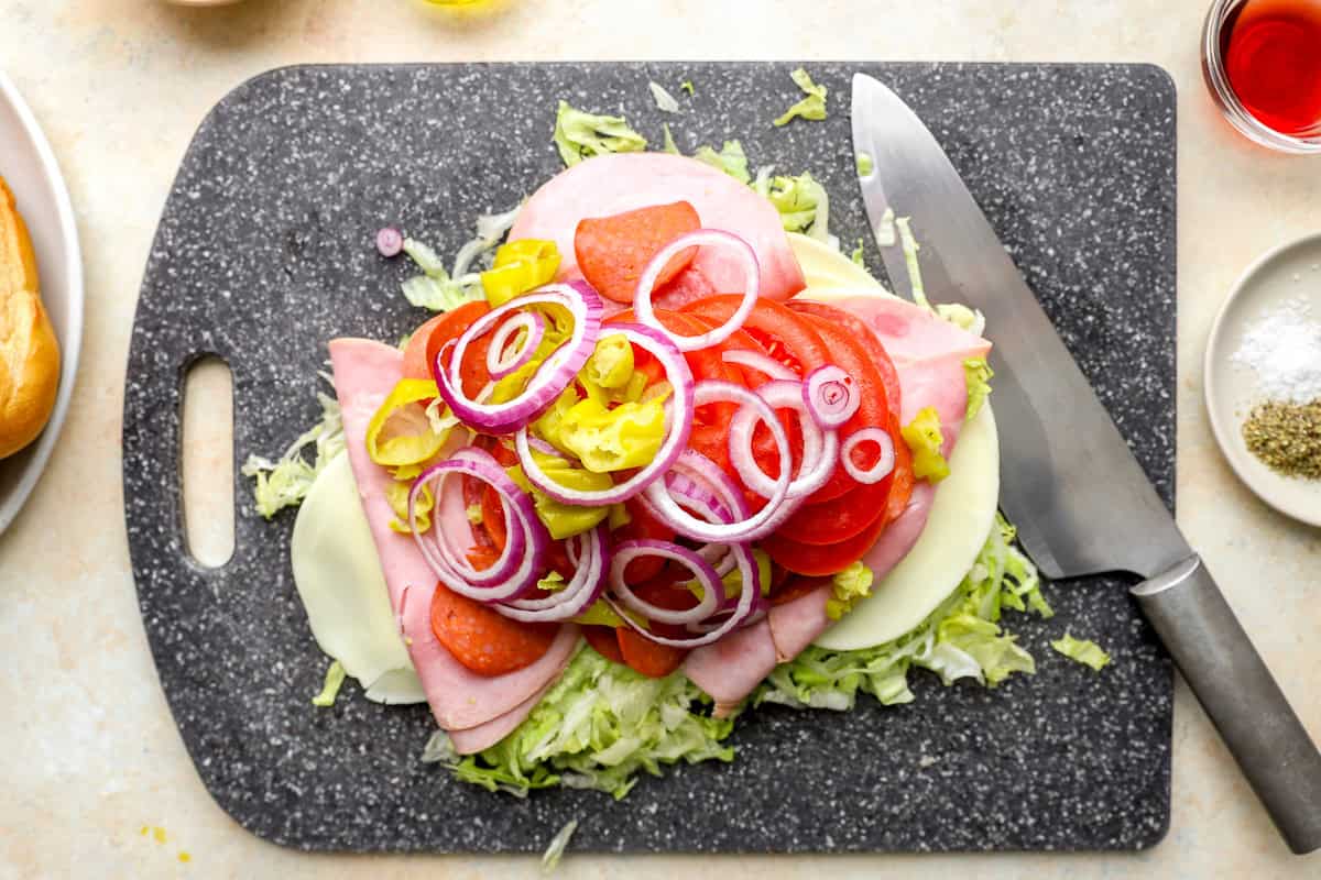 Lettuce, salami, ham, pepperoni, provolone, tomato slices, red onion, and pepperoncini layered on a cutting board before chopping.
