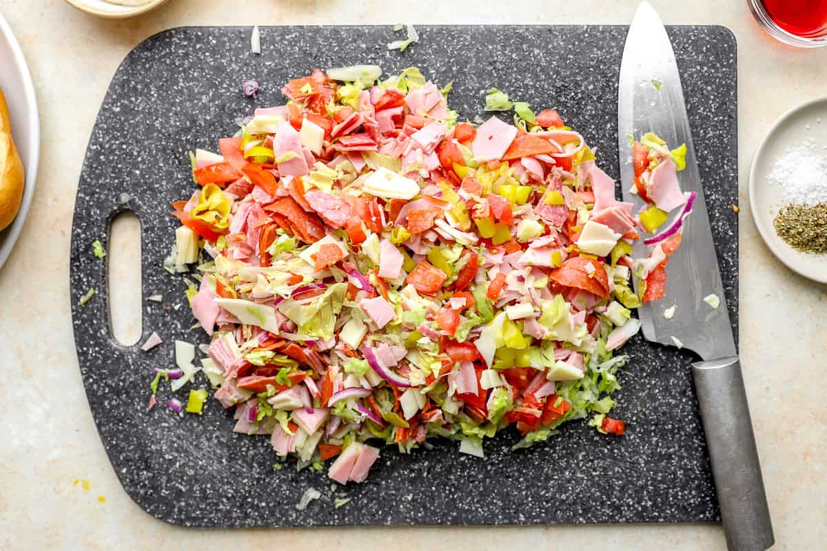 Chopped Italian sandwich ingredients being finely chopped together on a cutting board with a chef’s knife.