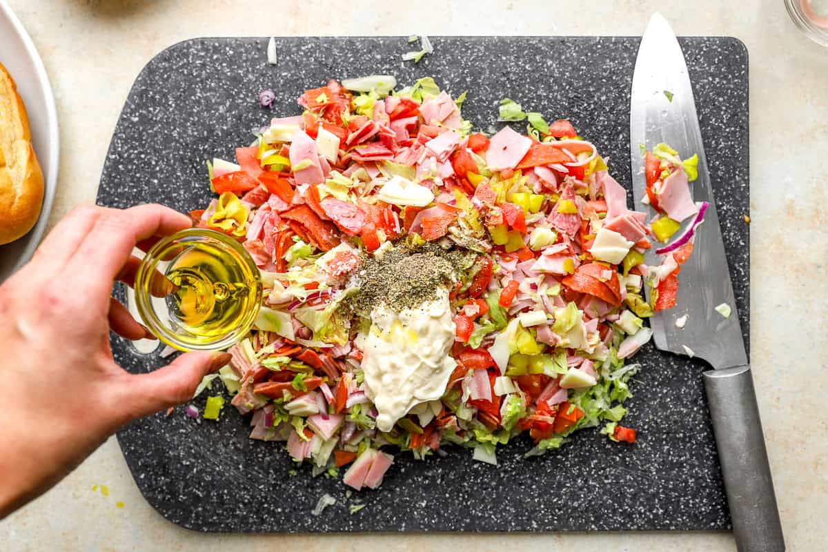 Mayonnaise, olive oil, red wine vinegar, and Italian seasoning added to chopped Italian sandwich ingredients on a cutting board.