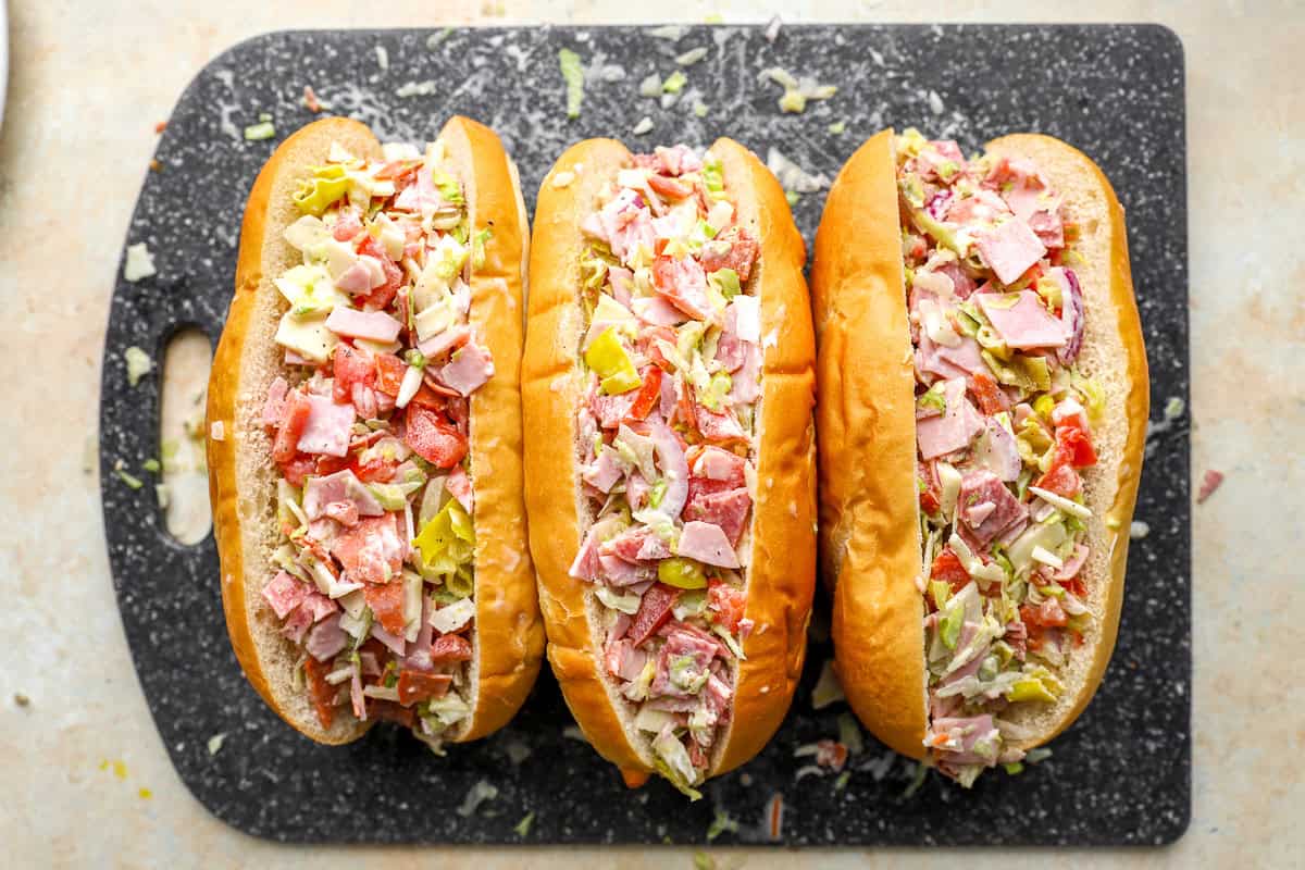 Three chopped Italian sandwiches assembled in hoagie rolls lined up on a cutting board.
