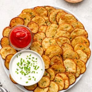 Crispy oven baked cottage fries arranged on a platter with ketchup and creamy dipping sauce, garnished with chopped herbs.