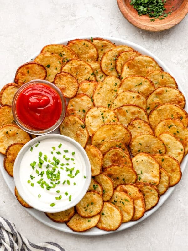 Crispy oven baked cottage fries arranged on a platter with ketchup and creamy dipping sauce, garnished with chopped herbs.