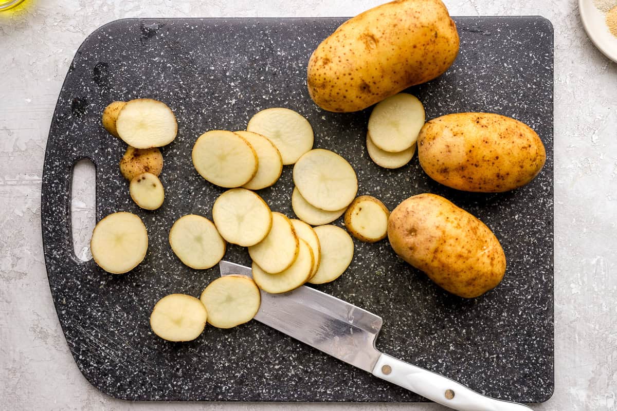 Russet potatoes sliced into even ¼-inch rounds on a cutting board with a knife.