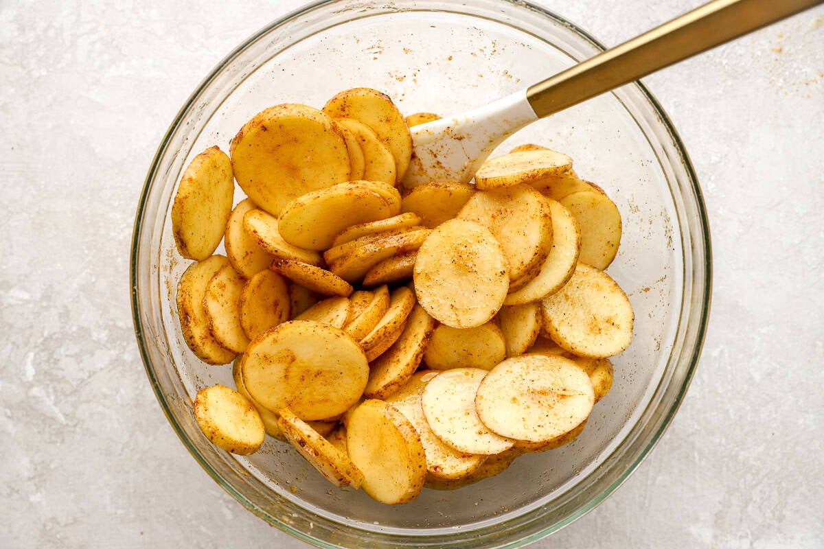 Potato slices tossed with olive oil and seasonings in a mixing bowl before baking.