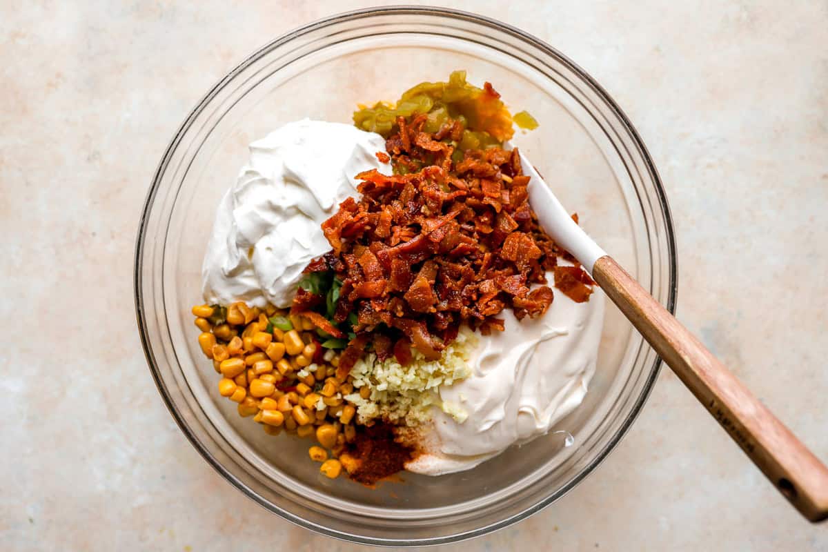 ngredients for crack corn dip in a mixing bowl, including bacon, corn, sour cream, mayonnaise, green chilies, garlic, and spices.