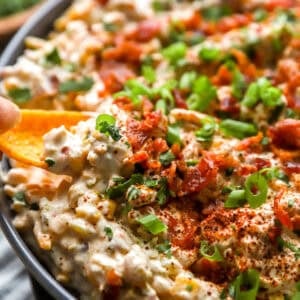 Crack corn dip topped with bacon, green onions, and seasoning, served in a bowl with a Fritos Scoop dipping in.
