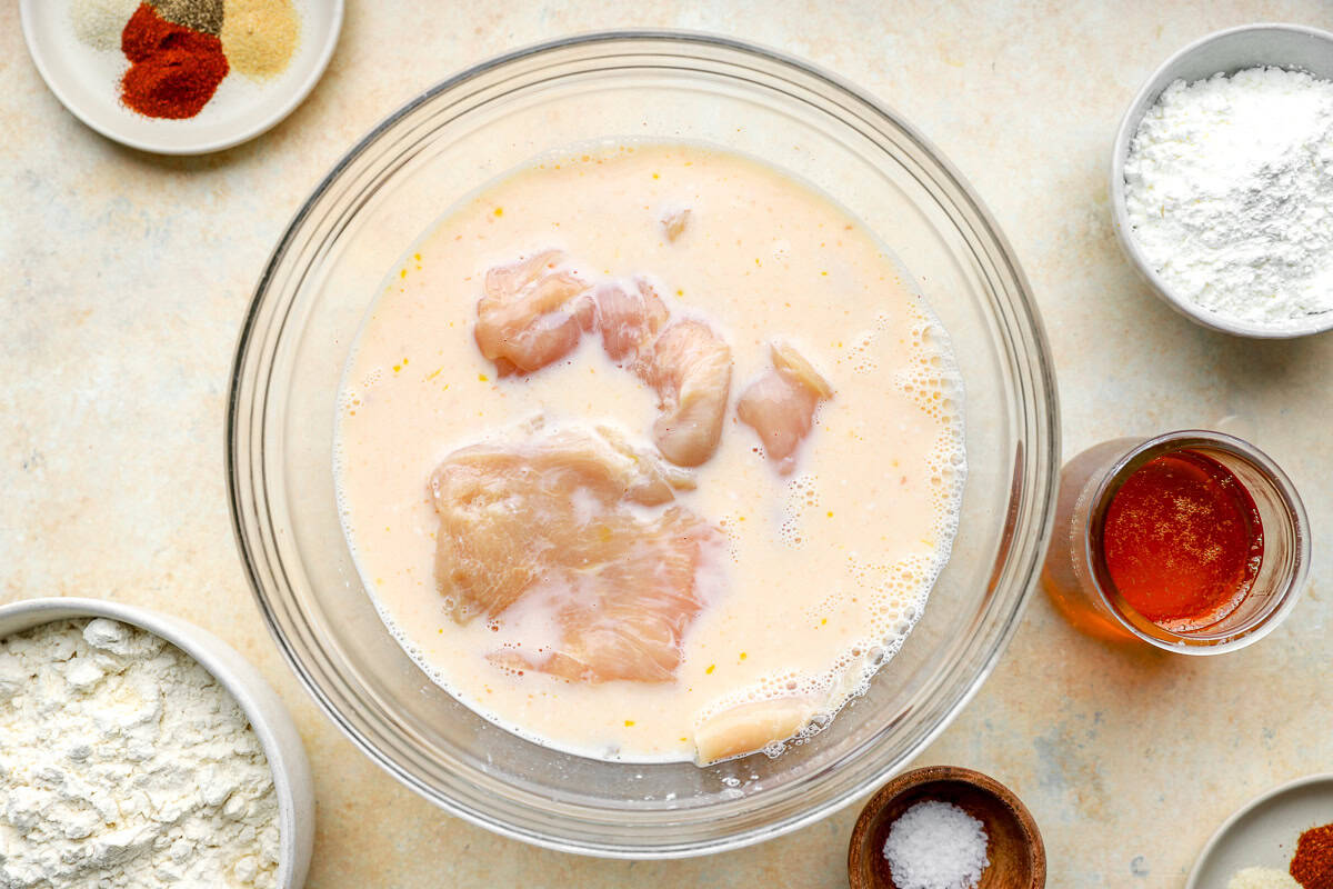 Raw chicken pieces marinating in a bowl of buttermilk, pickle brine, and hot sauce.