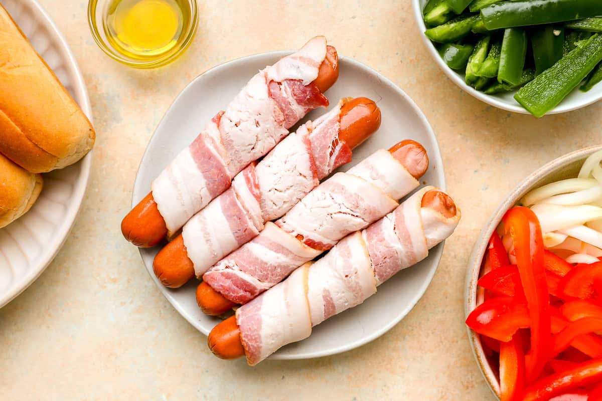 Raw hot dogs wrapped in bacon on a plate with sliced peppers, onions, jalapeños, olive oil, and buns prepped for Mexican hot dogs.