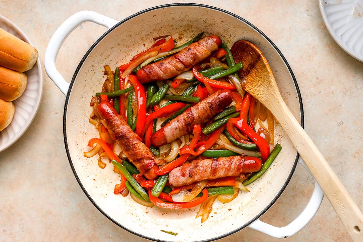 Bacon-wrapped hot dogs cooking in a skillet with sautéed peppers and onions.