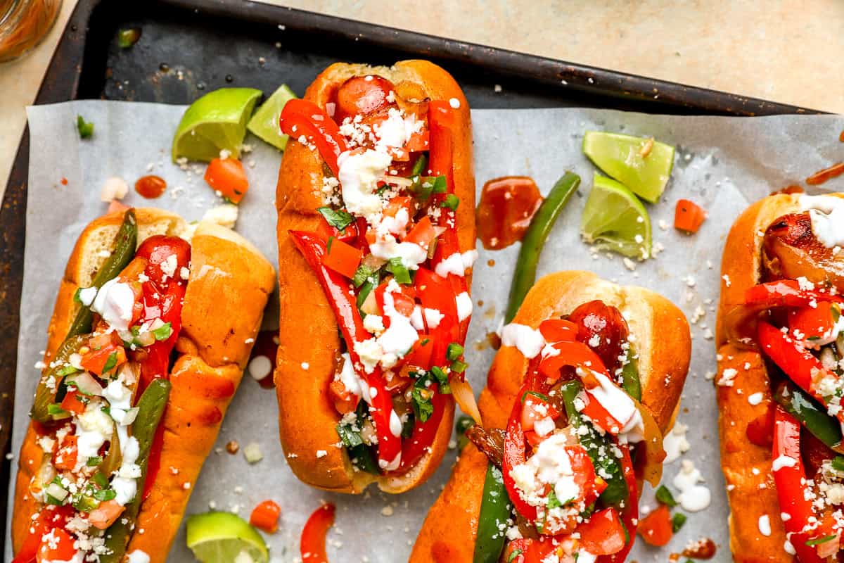 Overhead view of bacon-wrapped Mexican hot dogs with peppers, pico de gallo, cheese, and lime wedges.