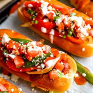 Bacon-wrapped Mexican hot dogs topped with peppers, jalapeños, pico de gallo, cotija cheese, and sour cream in toasted buns.