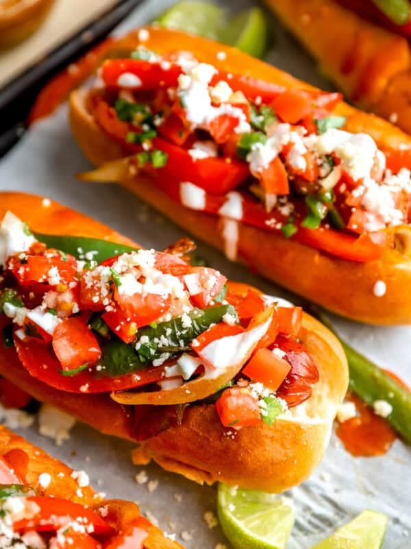 Bacon-wrapped Mexican hot dogs topped with peppers, jalapeños, pico de gallo, cotija cheese, and sour cream in toasted buns.