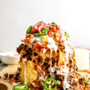 Tall trash can nachos stacked with tortilla chips, seasoned beef, black beans, melted cheese, sour cream, jalapeños, pico de gallo, and pickled onions