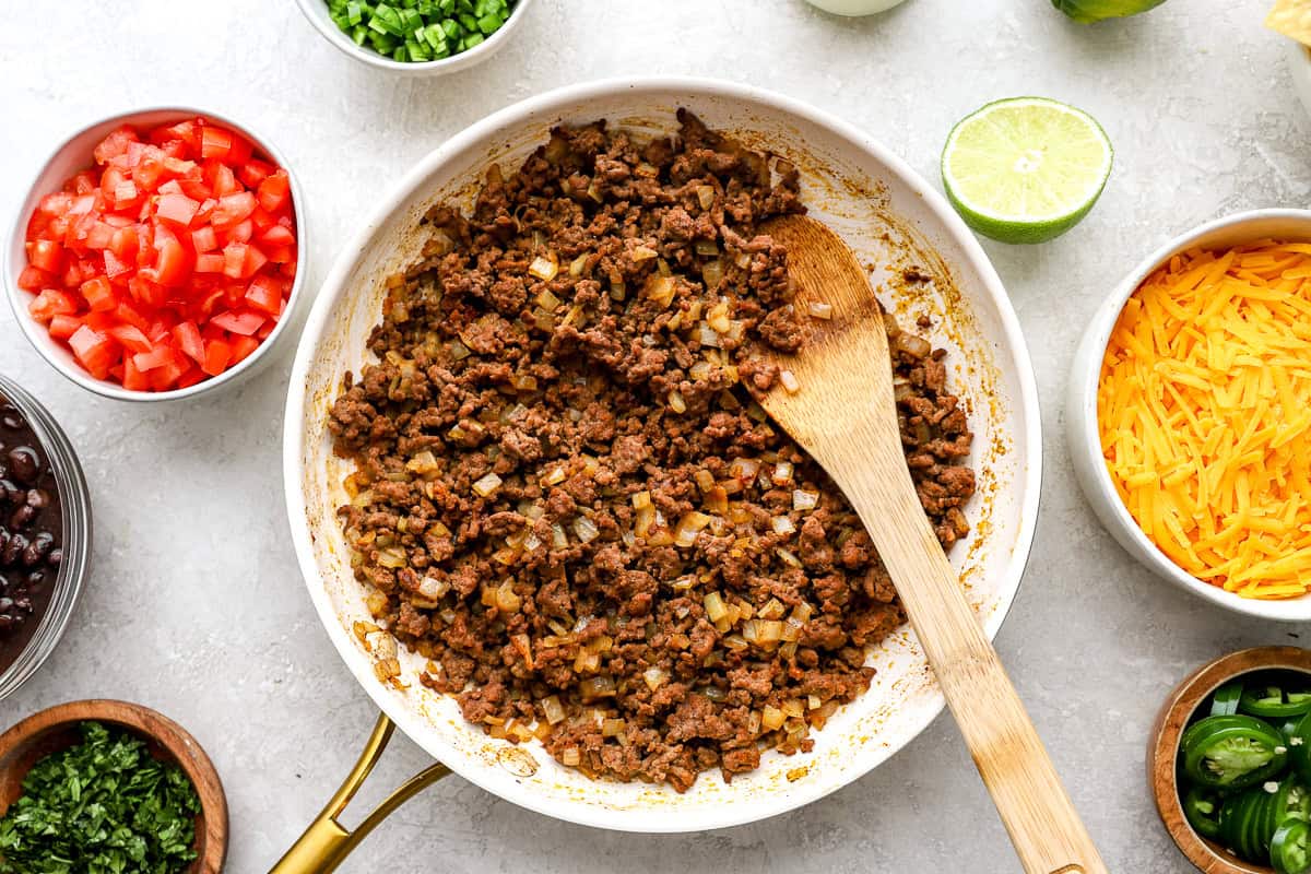 Ground beef cooked with chopped onion in a skillet for trash can nachos, surrounded by prepared toppings