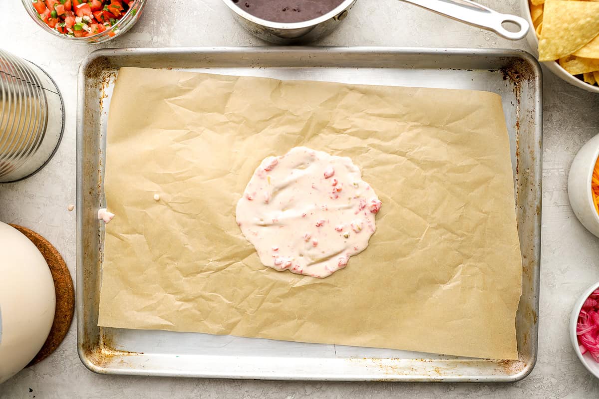 Parchment-lined sheet pan with a round layer of queso sauce for building trash can nachos