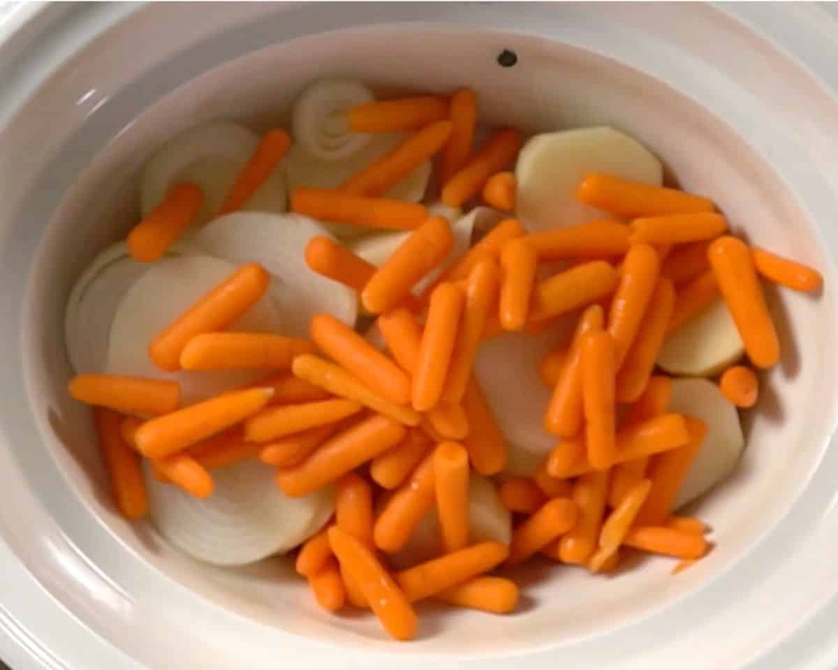Sliced onions, baby carrots, and halved red potatoes layered in the bottom of a slow cooker for corned beef and cabbage