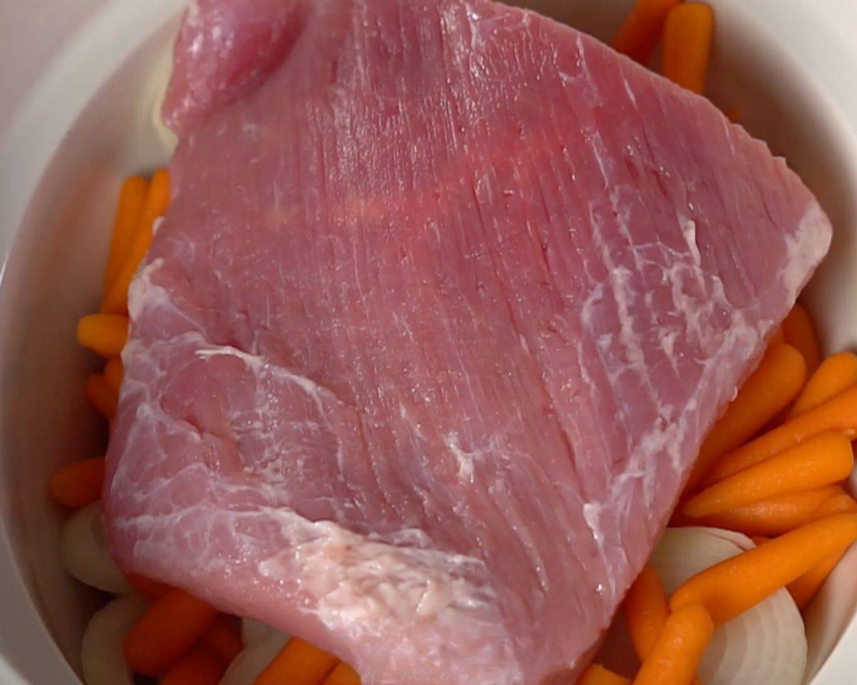 Raw corned beef brisket placed on top of carrots, potatoes, and onions in slow cooker before cooking