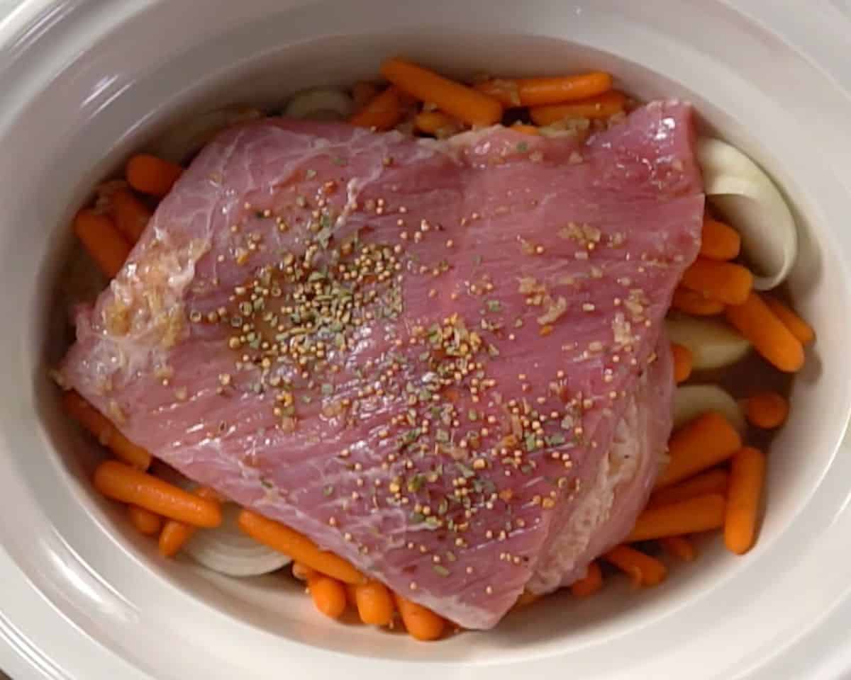 Corned beef brisket topped with spice packet seasoning in slow cooker over vegetables before cooking