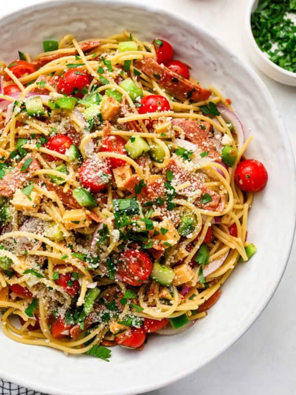 Spaghetti salad with Italian dressing, cheddar cubes, pepperoni, tomatoes, cucumber, red onion, parsley, and parmesan in a large bowl