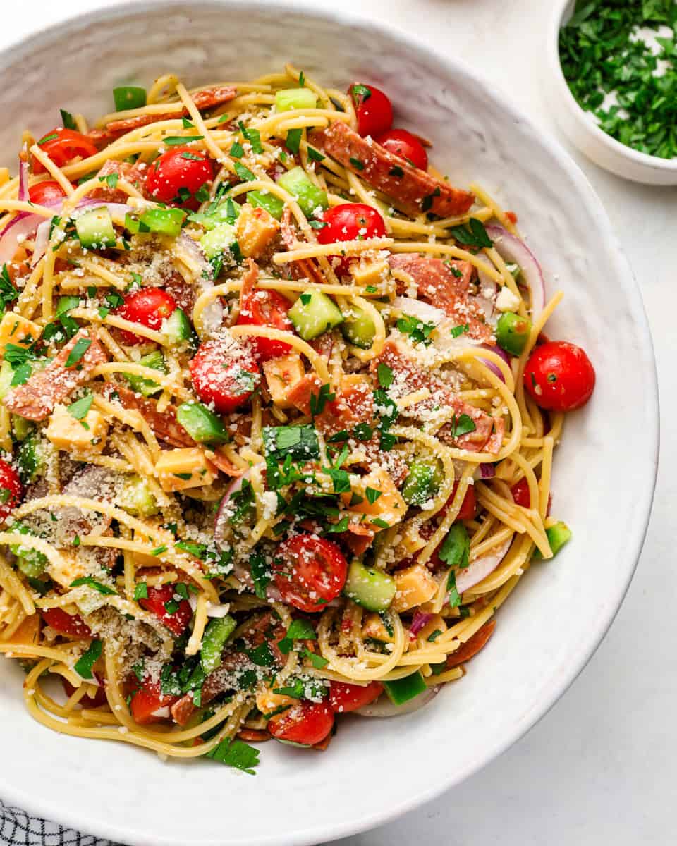 Spaghetti salad with Italian dressing, cheddar cubes, pepperoni, tomatoes, cucumber, red onion, parsley, and parmesan in a large bowl