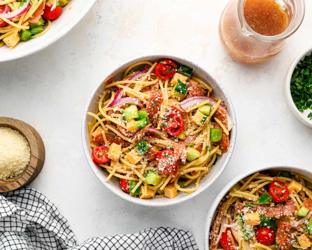 Individual bowls of spaghetti salad served with parmesan, parsley, and Italian dressing nearby