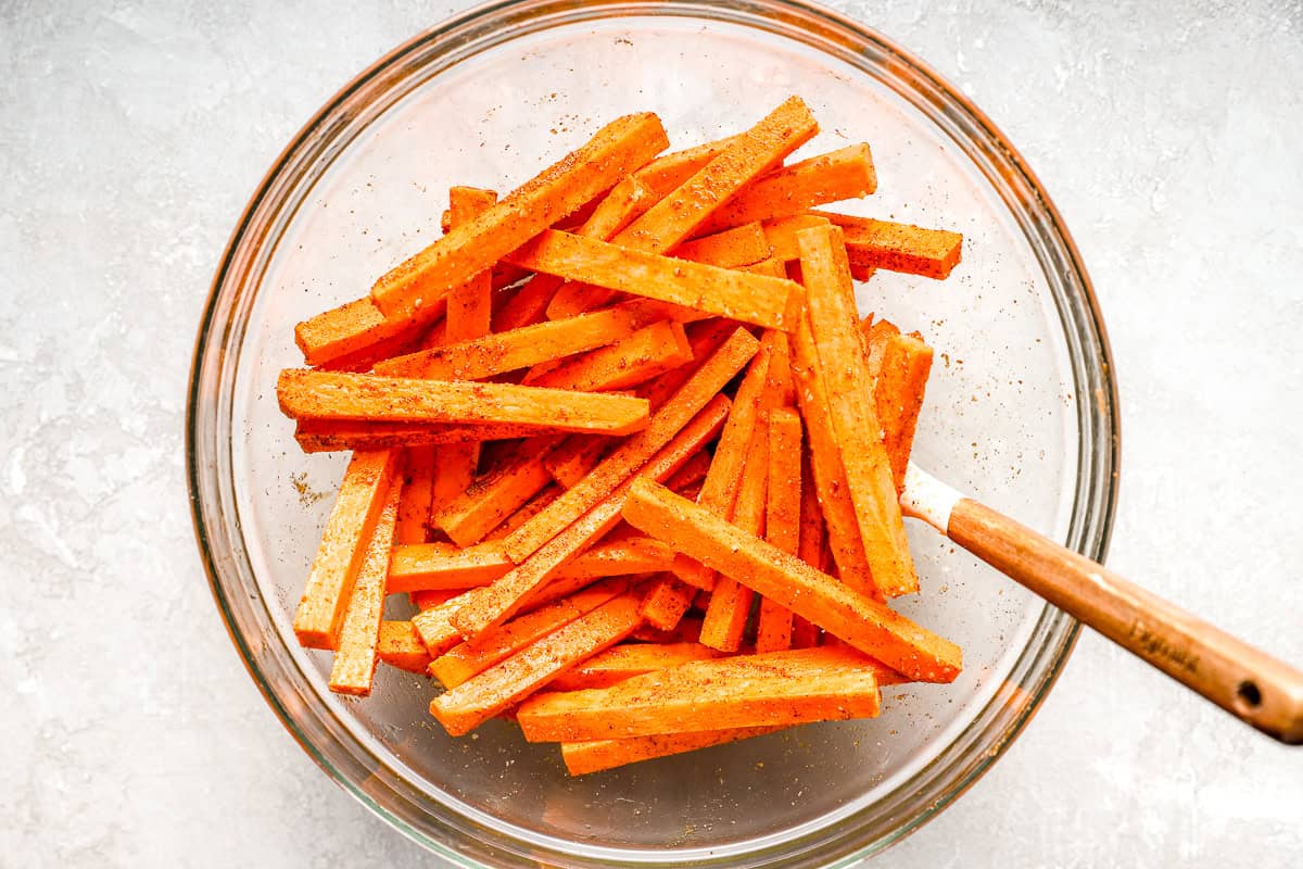 Raw sweet potato fries in a glass bowl tossed with oil and seasoning before air frying.