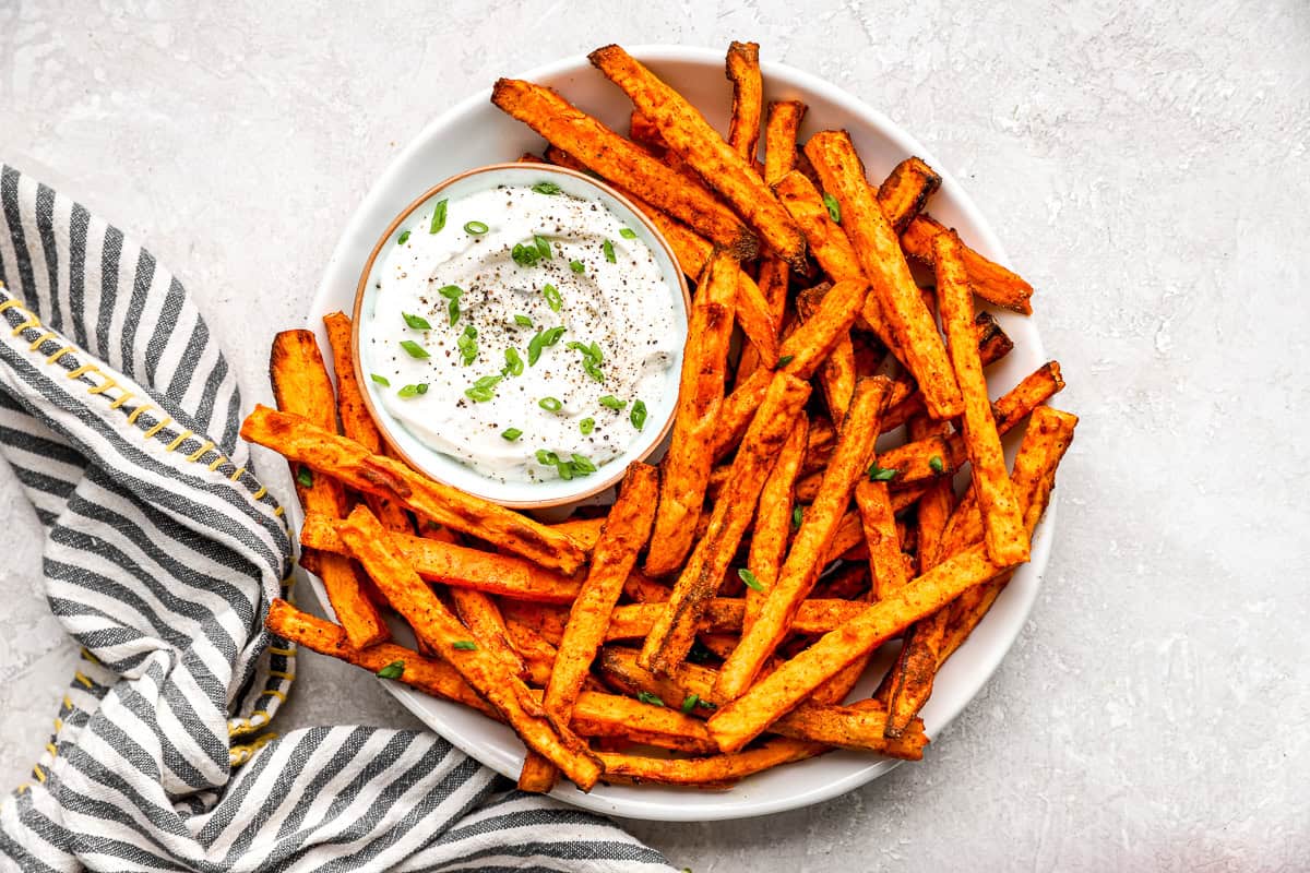 Overhead view of crispy air fryer sweet potato fries with dipping sauce on a white platter and striped towel.