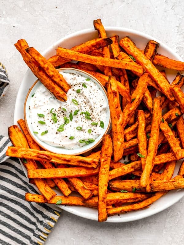 Air fryer sweet potato fries served on a platter with creamy dipping sauce, garnished with green onions and cracked pepper.