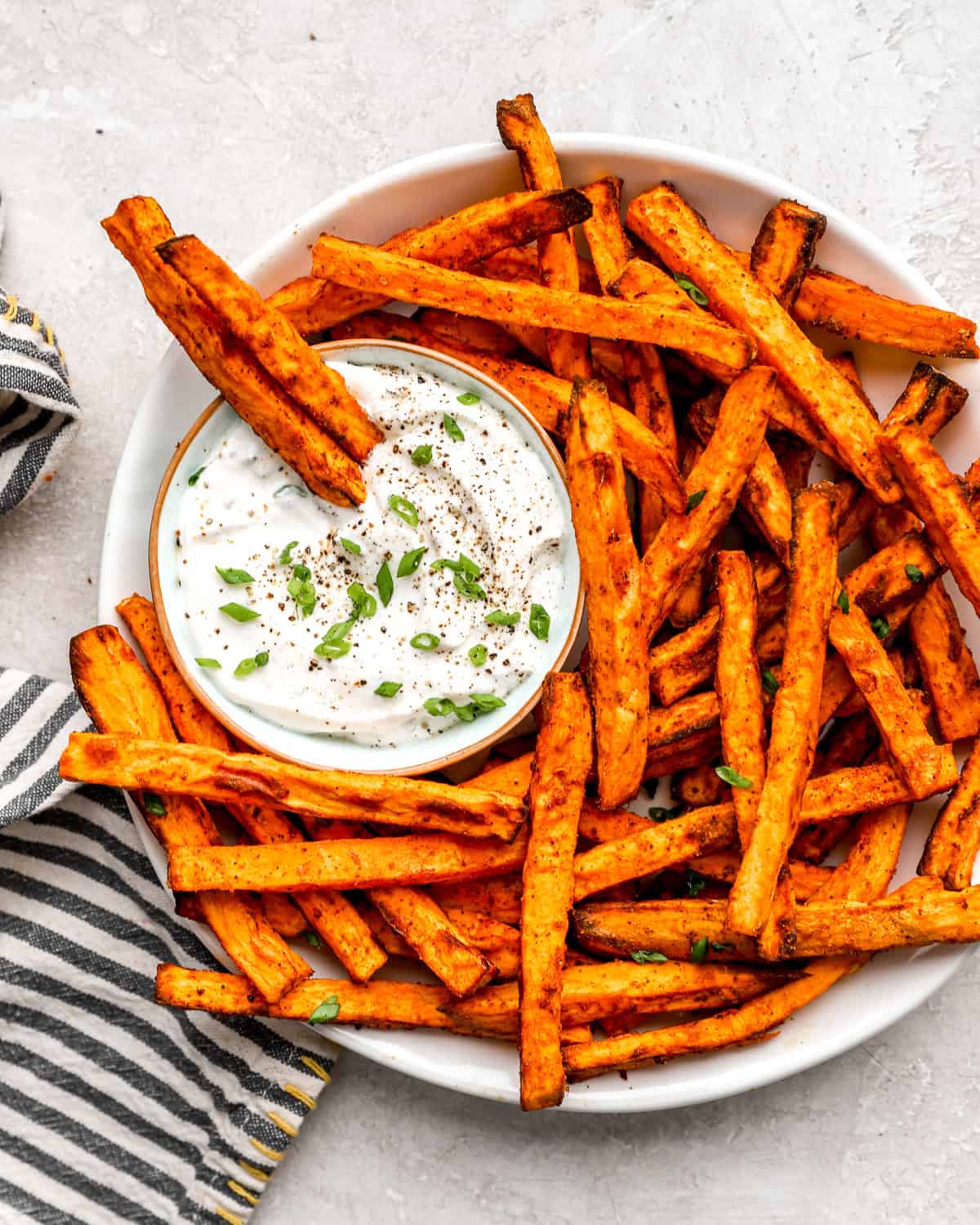 Air fryer sweet potato fries served on a platter with creamy dipping sauce, garnished with green onions and cracked pepper.