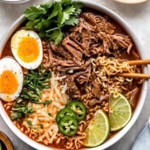 Birria ramen bowl topped with shredded beef, jammy soft boiled eggs, jalapeño slices, green onions, cilantro, shredded cheese, and lime wedges.