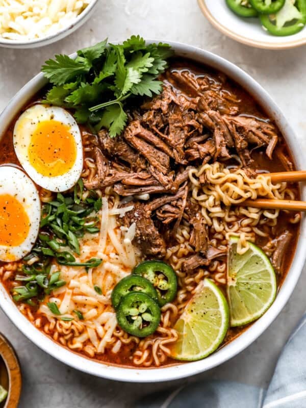 Birria ramen bowl topped with shredded beef, jammy soft boiled eggs, jalapeño slices, green onions, cilantro, shredded cheese, and lime wedges.