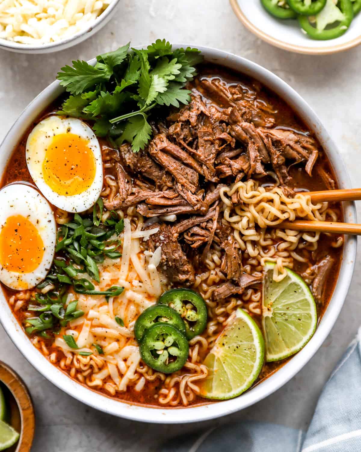 Birria ramen bowl topped with shredded beef, jammy soft boiled eggs, jalapeño slices, green onions, cilantro, shredded cheese, and lime wedges.