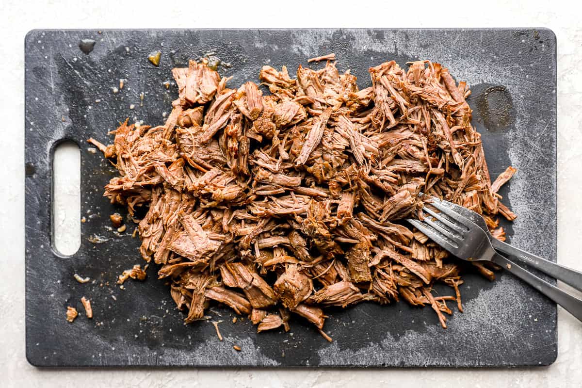 Shredded beef birria on a cutting board with a fork, showing tender, juicy texture after slow cooking.
