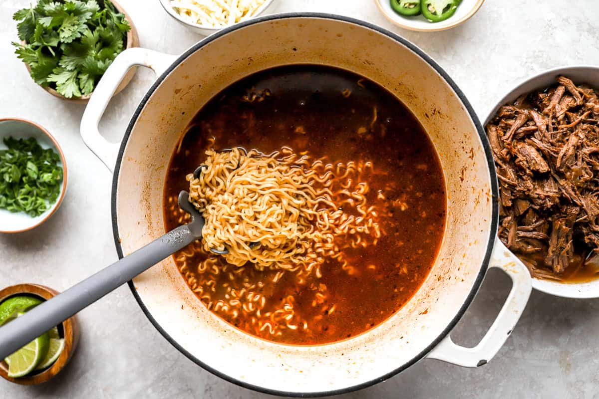 Ramen noodles cooking in rich red birria consommé inside a large Dutch oven with beef and toppings nearby.