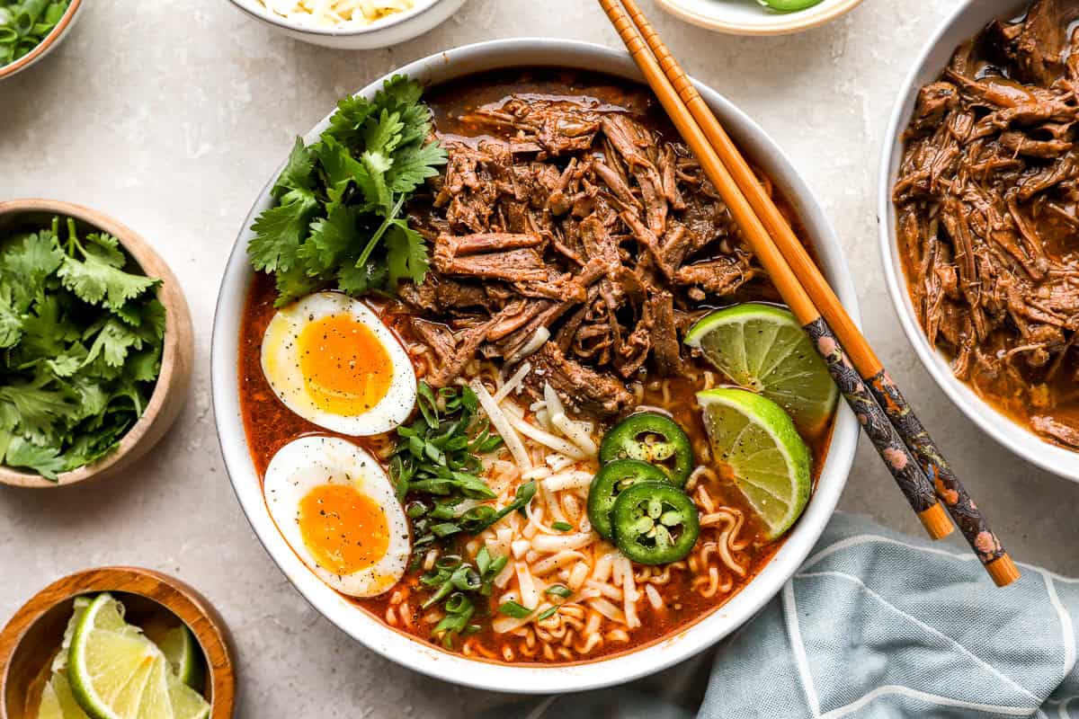 Birria ramen bowl with shredded beef, soft boiled eggs, fresh cilantro, jalapeños, lime wedges, and chopsticks.