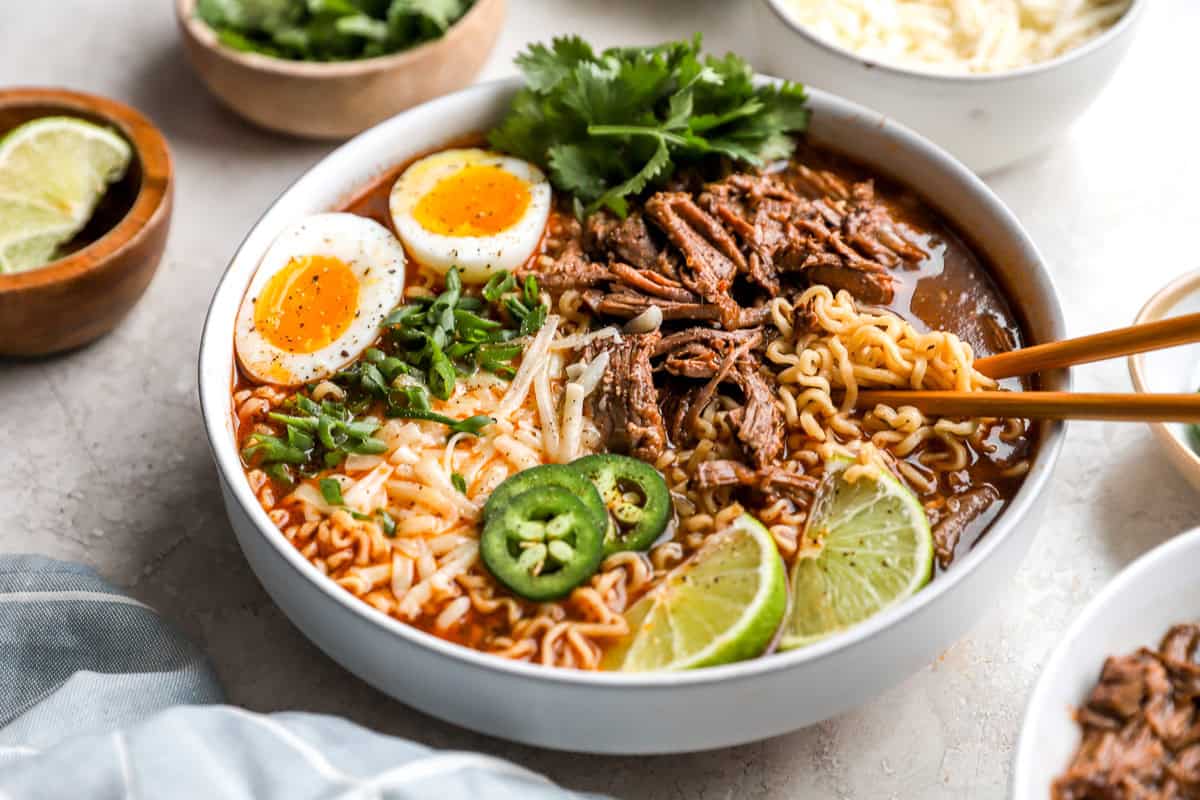 Close-up of birria ramen with tender shredded beef, ramen noodles, jammy eggs, green onions, and melted cheese in rich broth.