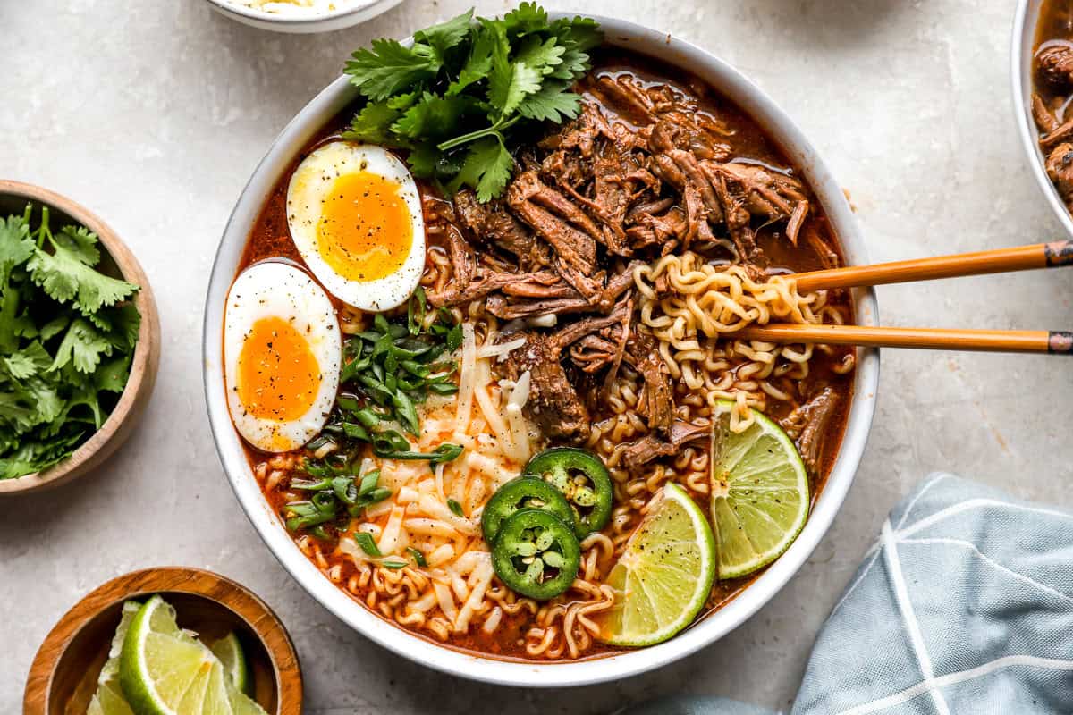 Overhead view of birria ramen bowl with beef, noodles, eggs, cilantro, jalapeños, and lime wedges on a light countertop.