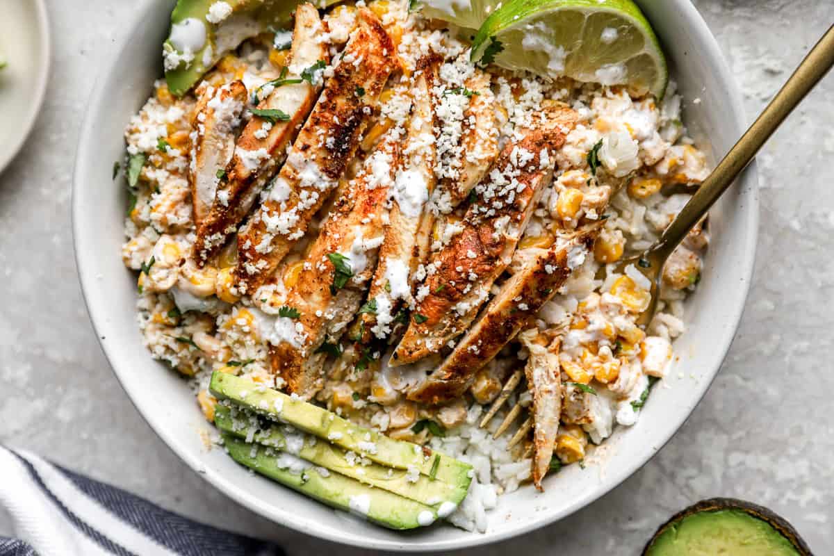 Close-up of sliced grilled chicken over creamy street corn and rice, topped with cotija and lime.