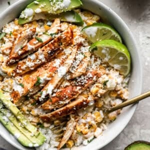 Street corn chicken bowl with sliced grilled chicken, creamy corn mixture, white rice, avocado slices, cotija cheese, and lime wedges.
